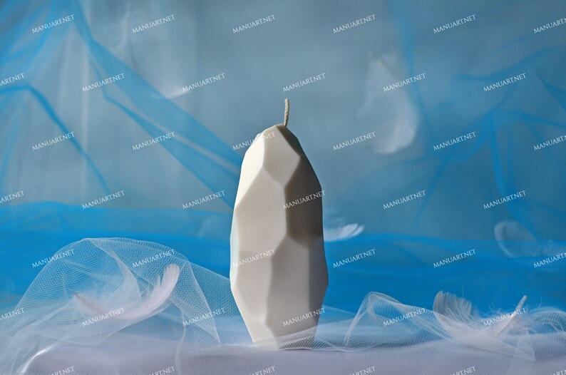 May include: A white geometric candle with a pointed top sits on a blue and white sheer fabric background.