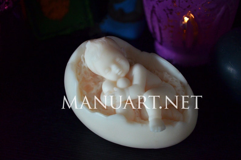 May include: A white baby figurine sleeping inside a cracked egg shell. The baby is holding a small object in its hand. The figurine is made of a smooth, white material.