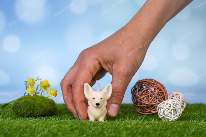 May include: A hand holds a small white candle shaped like a corgi dog. The candle is sitting on a bed of green grass with brown and white woven balls nearby.