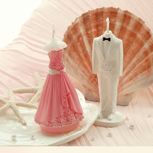 May include: A pair of wedding cake toppers, one in a white tuxedo and the other in a pink dress. The toppers are made of white and pink wax and are standing on a white base.