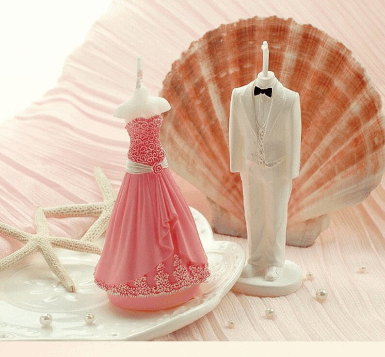 May include: A pair of wedding cake toppers, one in a white tuxedo and the other in a pink dress. The toppers are made of white and pink wax and are standing on a white base.
