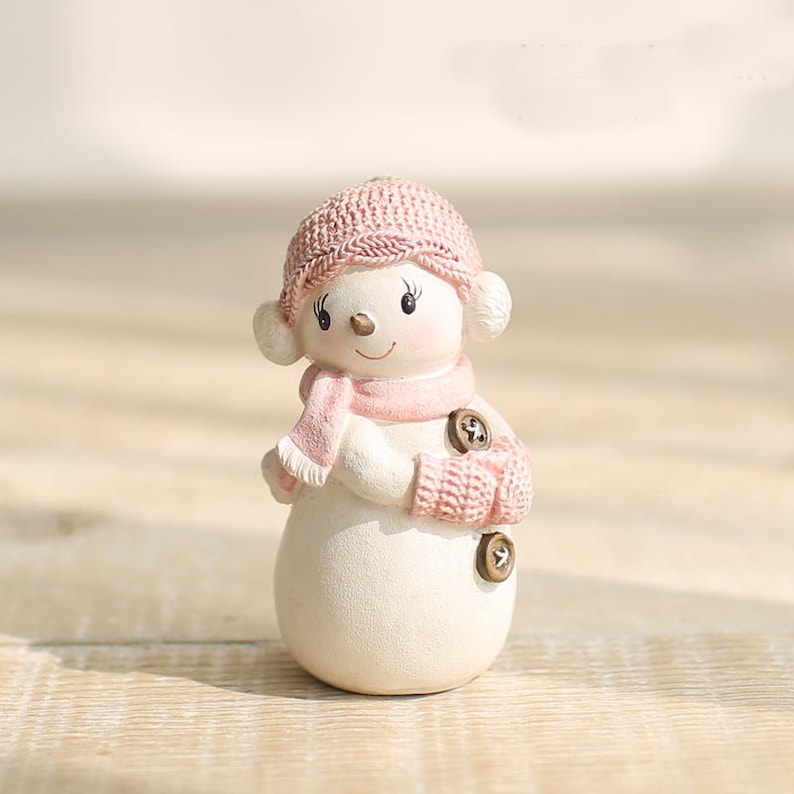 May include: A small white snowman figurine with a pink knitted hat and scarf. The snowman has black eyes and a small smile.