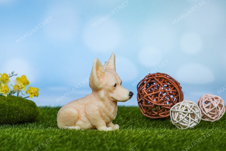 May include: A cream-colored ceramic figurine of a corgi dog sitting on a green artificial grass surface. The dog is looking to the right. There are three brown and white woven balls in the background.