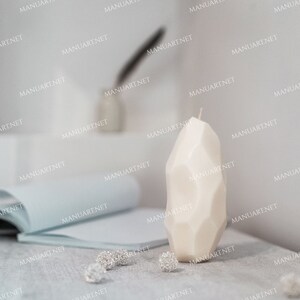 May include: A white, geometrically shaped candle on a light gray surface. The candle is tall and narrow, with a pointed top. There are small, silver decorative items scattered around the candle.
