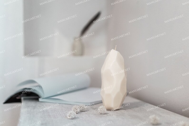 May include: A white, geometrically shaped candle on a light gray surface. The candle is tall and narrow, with a pointed top. There are small, silver decorative items scattered around the candle.