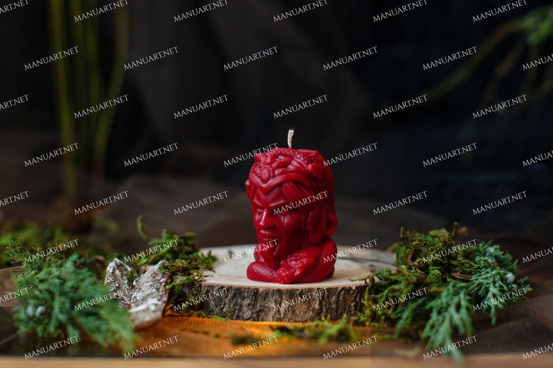 Pu&ograve; includere: Una candela rossa a forma di testa umana con un viso dettagliato e uno stoppino in cima. La candela &egrave; appoggiata su un disco di legno con muschio verde intorno.