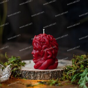 Pu&ograve; includere: Una candela rossa a forma di testa di donna con serpenti intrecciati nei capelli. La candela &egrave; appoggiata su una superficie di legno ricoperta di muschio.