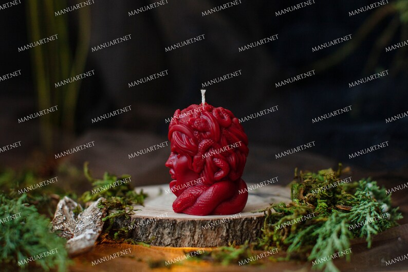 Pu&ograve; includere: Una candela rossa a forma di testa di donna con serpenti intrecciati nei capelli. La candela &egrave; appoggiata su una superficie di legno ricoperta di muschio.