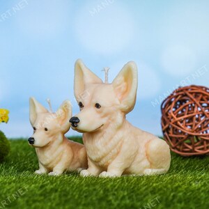 May include: Two beige candle sculptures of corgi dogs, one larger than the other, sitting on a green grassy surface.