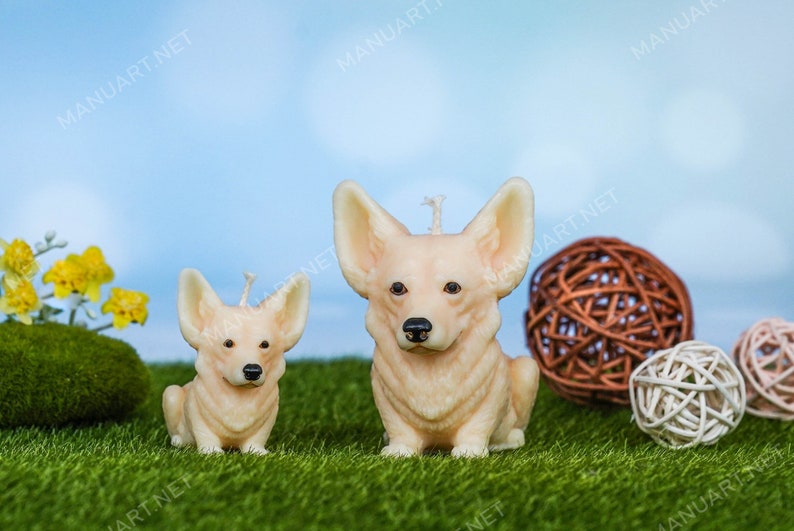 May include: Two realistic-looking, cream-colored, candle sculptures of corgi dogs. The larger corgi is sitting with its paws forward, while the smaller corgi is sitting with its paws tucked under its body. Both candles have a small wick on top. The candles are on a green, artificial grass surface.