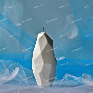 May include: A white geometric candle with a sharp, angular design. The candle is sitting on a blue and white sheer fabric background.