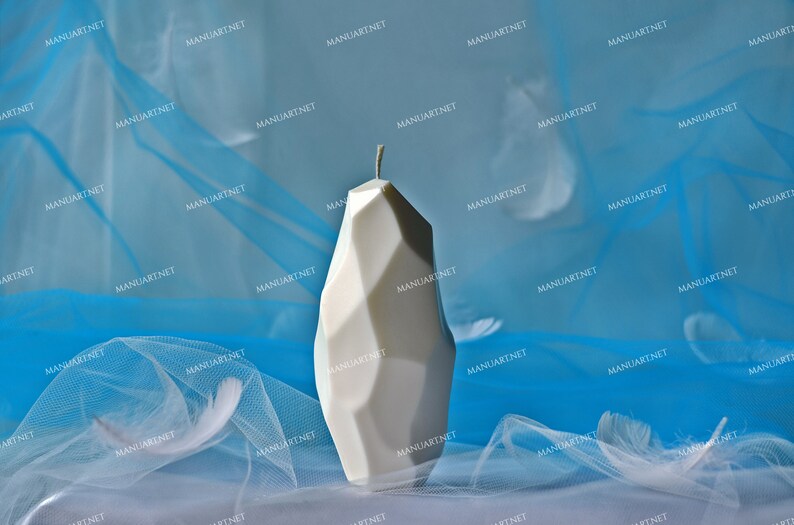 May include: A white geometric candle with a sharp, angular design. The candle is sitting on a blue and white sheer fabric background.