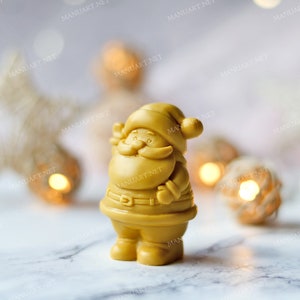 May include: A small, golden yellow Santa Claus figurine with a white beard and a red hat. The figurine is standing on a white surface with a blurred background of white and gold decorations.