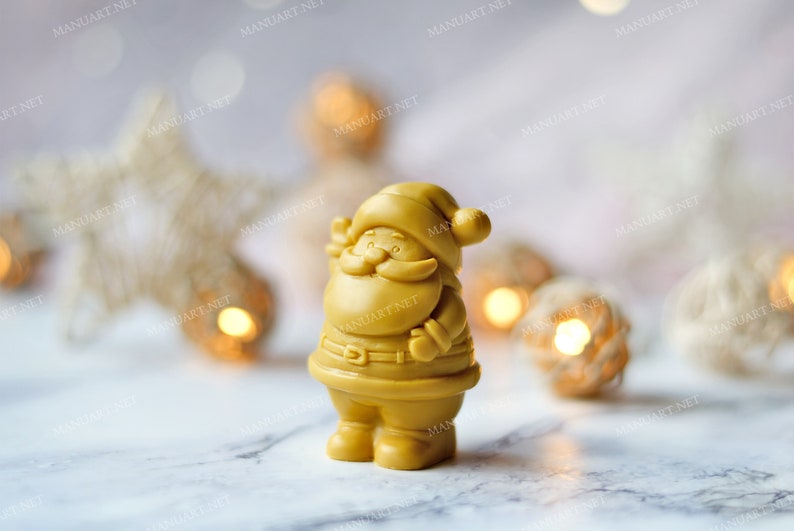 May include: A small, golden yellow Santa Claus figurine with a white beard and a red hat. The figurine is standing on a white surface with a blurred background of white and gold decorations.