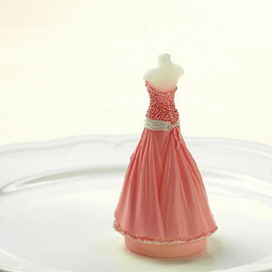 May include: A pink and white wedding cake topper featuring a bride in a pink gown with a white sash and a floral bodice.