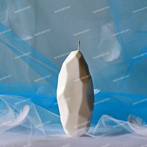 May include: A white, geometrically shaped candle with a pointed top sits on a white surface covered in a blue and white sheer fabric.