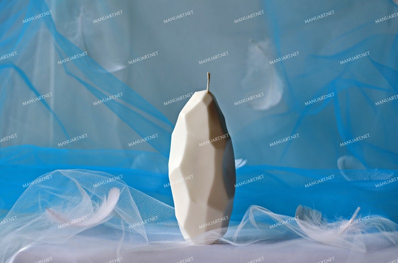 May include: A white, geometrically shaped candle with a pointed top sits on a white surface covered in a blue and white sheer fabric.