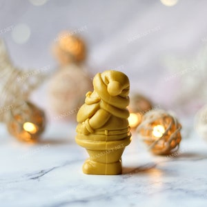 May include: A small, golden yellow Santa Claus figurine made of wax or soap. The figurine is standing on a white marble surface with a blurred background of white and gold string lights.