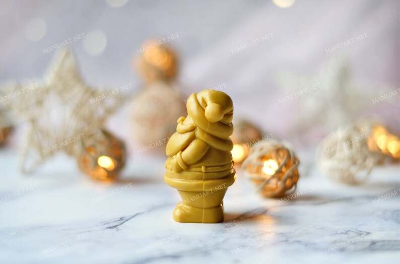 May include: A small, golden yellow Santa Claus figurine made of wax or soap. The figurine is standing on a white marble surface with a blurred background of white and gold string lights.