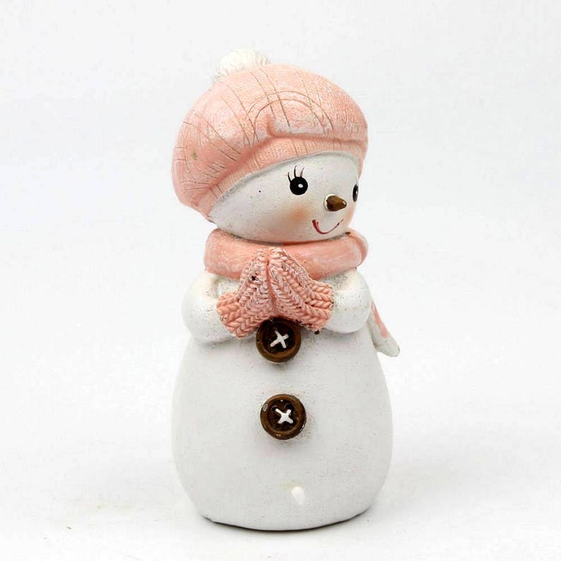 May include: A small pink snowman figurine with a pink hat and mittens. The snowman has brown button eyes and a brown button nose.
