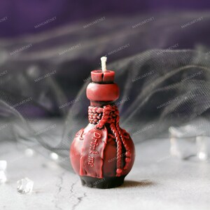 Pode incluir: Uma vela de garrafa de vidro vermelha com um design de corda e o texto "Drink me" no lado. A vela est&aacute; sobre uma superf&iacute;cie branca com um tecido preto transparente no fundo.