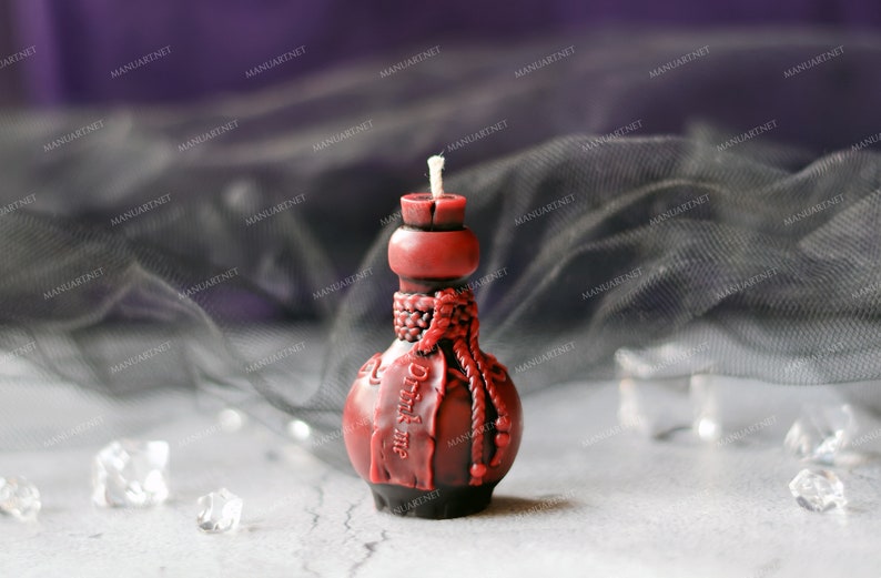 Pode incluir: Uma vela de garrafa de vidro vermelha com um design de corda e o texto "Drink me" no lado. A vela est&aacute; sobre uma superf&iacute;cie branca com um tecido preto transparente no fundo.