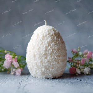 May include: A white egg-shaped candle with a floral design. The candle is surrounded by pink flowers.