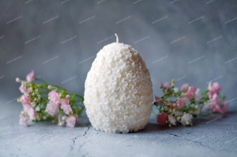 May include: A white egg-shaped candle with a floral design. The candle is surrounded by pink flowers.