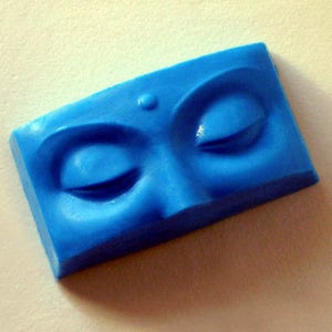 May include: A blue plastic mold of a face with closed eyes and a dot on the forehead. The mold is a rectangular shape.