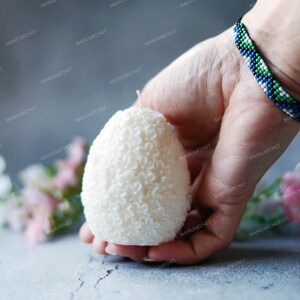 May include: A white egg-shaped candle with a floral design. The candle is being held in a hand with a beaded bracelet.
