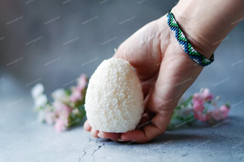 May include: A white egg-shaped candle with a floral design. The candle is being held in a hand with a beaded bracelet.