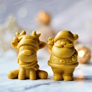 May include: Two small, golden yellow, Christmas-themed figurines. A reindeer with a red and white hat and a Santa Claus with a white beard and a red belt.