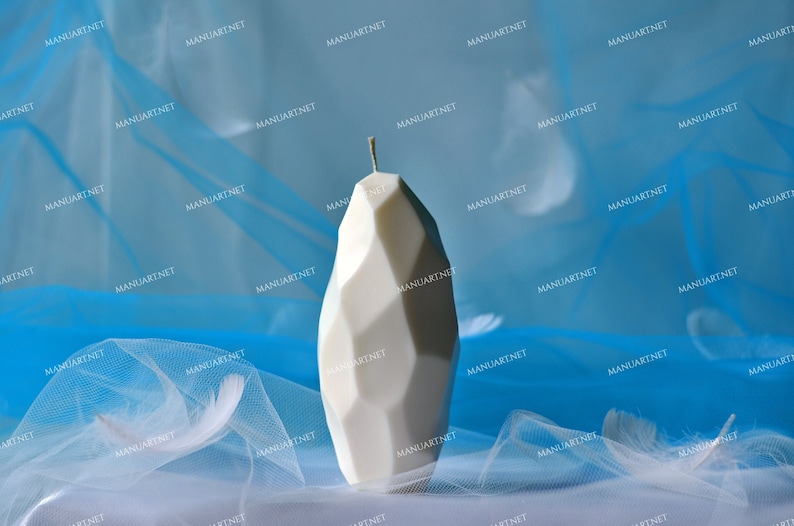 May include: A white, geometrically shaped candle with a pointed top. The candle is sitting on a white surface with a blue sheer fabric behind it.