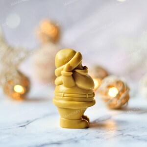 May include: A golden yellow Santa Claus figurine with a white hat and a red scarf. The figurine is standing on a white marble surface with a blurred background of white and gold decorations.