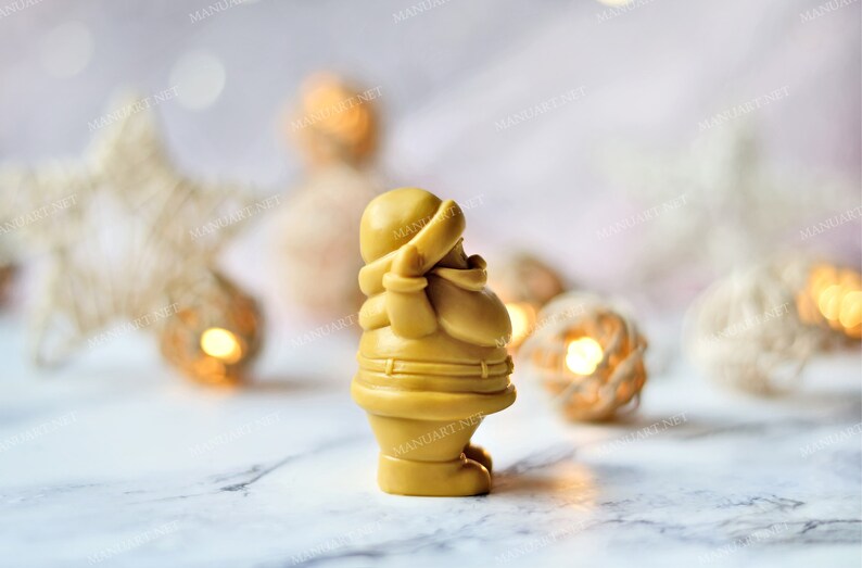 May include: A golden yellow Santa Claus figurine with a white hat and a red scarf. The figurine is standing on a white marble surface with a blurred background of white and gold decorations.