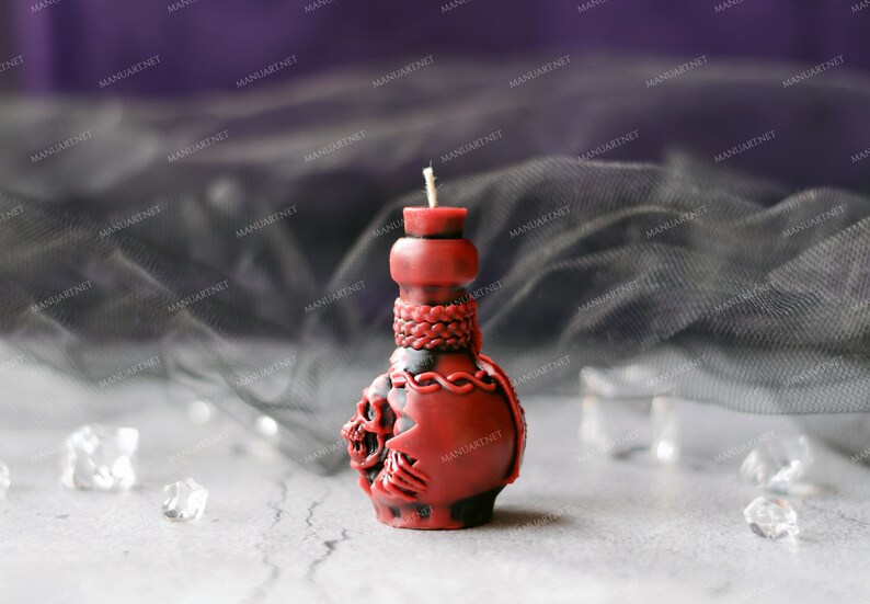 Pode incluir: Uma vela vermelha em forma de caveira com um design de corda preta e vermelha. A vela est&aacute; sobre uma superf&iacute;cie cinza com um tecido preto transparente no fundo.