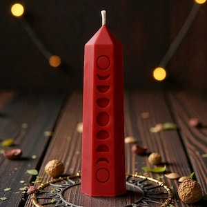 Può includere: Una candela a colonna rossa, alta, con un design geometrico e uno stoppino bianco. La candela presenta simboli delle fasi lunari in rilievo. È esposta su una superficie di legno con elementi decorativi.
