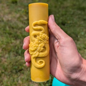 Op de afbeelding: Een gele cilindrische kaars met een gesneden ontwerp van een slang en bloemen. De kaars wordt in een hand gehouden tegen een vage groene achtergrond. Het slang- en bloemontwerp is een lichtere tint geel.