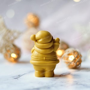 May include: A yellow, hand-crafted Santa Claus figurine with a white beard and a red hat. The figurine is standing on a white surface with a blurred background of white and gold decorations.