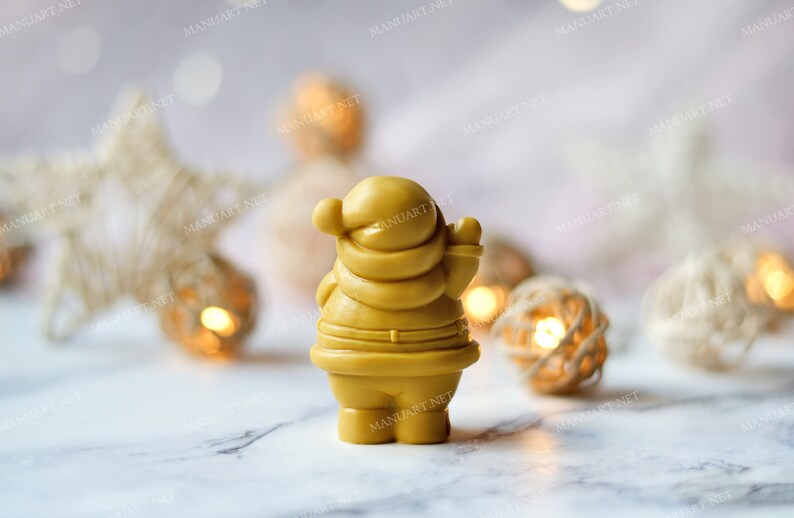 May include: A yellow, hand-crafted Santa Claus figurine with a white beard and a red hat. The figurine is standing on a white surface with a blurred background of white and gold decorations.