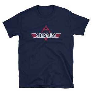 Puede incluir: Camiseta azul marino con la frase "STOP GUNS" en blanco, con efecto desgastado. El diseño incluye dos rifles cruzados y una estrella roja. La camiseta tiene cuello redondo y mangas cortas.