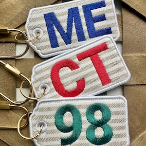 May include: Three embroidered keychains with gold-colored clasps. The top keychain reads "ME" in blue, the middle one reads "CT" in red, and the bottom one reads "98" in green. Each keychain has a beige and white striped background.