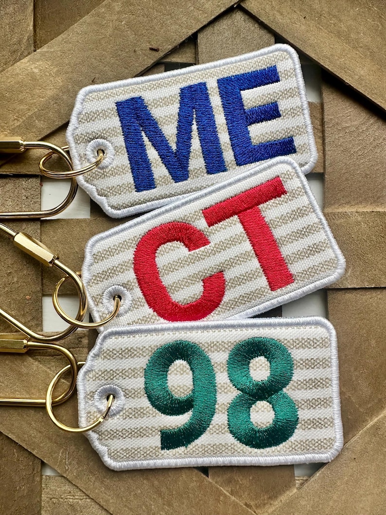 May include: Three embroidered keychains with gold-colored clasps. The top keychain reads "ME" in blue, the middle one reads "CT" in red, and the bottom one reads "98" in green. Each keychain has a beige and white striped background.