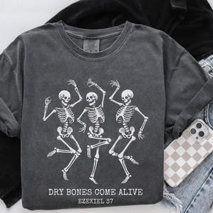 May include: A dark gray t-shirt with a graphic of three skeletons dancing. The text "Dry Bones Come Alive Ezekiel 37" is printed below the skeletons.