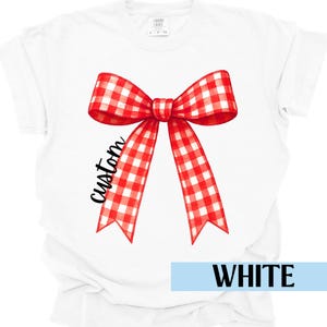 Puede incluir: Camiseta blanca con un gran diseño de lazo a cuadros rojo y blanco. La palabra "custom" está escrita en cursiva en la cinta del lazo. La palabra "WHITE" se muestra en un rectángulo azul claro en la parte inferior.