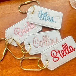 May include: White embroidered keychains with gold-colored clasps. The keychains feature names and titles in various colors: "Bride" in pink, "Mrs." in blue, "Collins" in pink, and "Stella" in red.