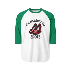 May include: White and green baseball tee with the text "IT'S ALL ABOUT THE SHOES" and an illustration of red shoes. The shirt has green sleeves and a green neckline.