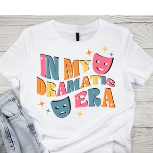 May include: White t-shirt with a colourful graphic design that says "In My Dramatic Era" with two colourful theatrical masks.