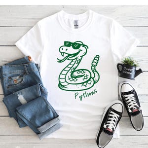 Könnte beinhalten: Weißes T-Shirt mit einer grünen Cartoon-Schlange mit Sonnenbrille und dem Wort "Pythons" in Schreibschrift. Das Shirt wird mit blauen Jeans und schwarz-weißen Turnschuhen auf einer hellen Holzmaserung präsentiert.