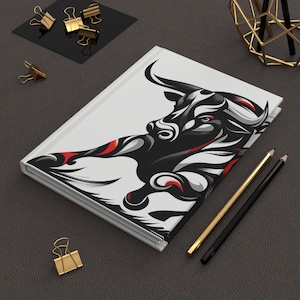Pode incluir: Um caderno branco com uma ilustração em preto e branco de um touro na capa. O touro está a carregar para a frente com os chifres apontados para a frente. A ilustração é estilizada e tem acentos vermelhos.